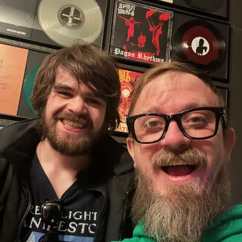Sam Pura and a friend laughing in front of the framed records, 2026
